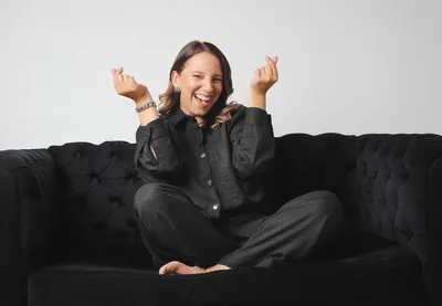 Retrato espontáneo en formato horizontal: niña riendo sentada sobre un sofá capitoné negro, con conjunto de mezclilla oscura y fondo blanco