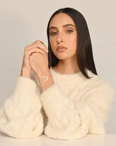 Fotografía de producto de joyería: manos entrelazadas lucen una pulsera dorada con dije de gato, modelo con suéter crema apoyada sobre mesa blanca