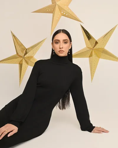 Editorial de temporada: modelo con vestido negro de cuello alto sentada en el piso, bajo estrellas doradas de papel suspendidas sobre fondo crema