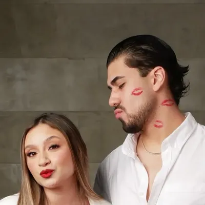 Sesión temática de pareja: ella con labial rojo junto a él con marcas de besos en el rostro y camisa blanca, sobre fondo de concreto gris